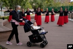 FILE - An elderly woman wearing a mask pushes a baby in a pram past plaza dancers at a park in Beijing, China, Thursday, Oct. 14, 2021. (AP Photo/Ng Han Guan)