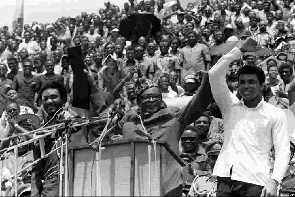 Le président du Zaïre Mobutu Sese Seko, lève la main de Muhammad Ali quand il le présente avec&nbsp; son challenger George Foreman avant le combat du championnat WBA/WBC au cours d&rsquo;un meeting populaire au stade du 20 Mai où les deux boxeurs disputeront&nbsp;le titre quelques jours plus tard. (AP Photo). 