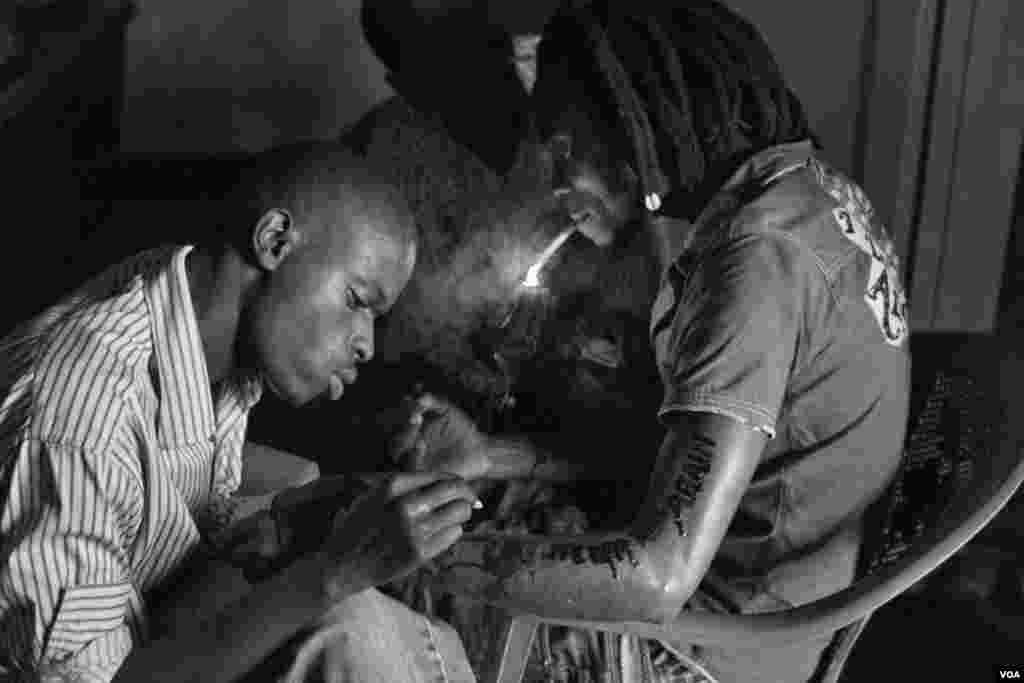 Negenzi Ali (left), age 23, makes a tattoo of a Rwandan genocide slogan &quot;never again&quot; on Tomy&#39;s arm. Negenzi Ali says &quot;I don&#39;t care about HIV transmission through my needles. Giporoso is killing our generation softly.&quot; Giporoso, Kigali, Rwanda, Nov. 5, 2013.