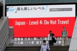 People wearing masks to help protect against the spread of the coronavirus walk in front of a screen showing the news on U.S. warning against visits to Japan, May 25, 2021, in Tokyo.