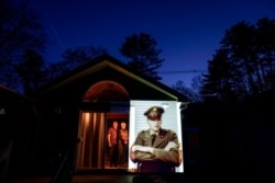 An image of veteran Roy Benson is projected onto the home of his daughter, Robin Benson Wilson, left, as she looks out a doorway with her husband, Donald, in Holland, Mass., May 13, 2020.