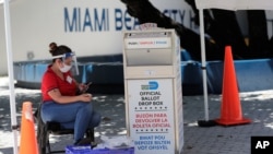Članica biračkog odbora nosi zaštitnu opremu dok posmatra glasačku kutiju sa glasovima pristiglim poštom, tokom ranog glasanja, 7. avgusta 2020, u Miami Beachu, Florida.