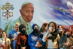 Para perempuan menunggu di luar Katedral Khaldea Saint Joseph, di Baghdad, Irak, Sabtu, 6 Maret 2021, di mana Paus Fransiskus, yang digambarkan pada poster raksasa di punggung mereka, sedang mengadakan misa. (Foto: AP)